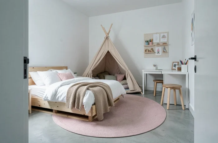 Chambre d'enfant style scandinave avec lit plateforme en bois, tipi en contreplaqué, bureau minimaliste et tapis rond rose blush sur béton poli